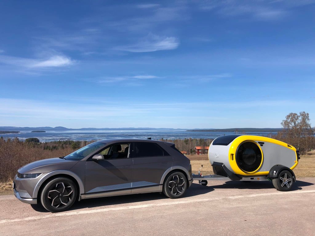 A MINK camper (teardrop caravan) parked by the side of a road, overlooking a large lake with mountains in the background.