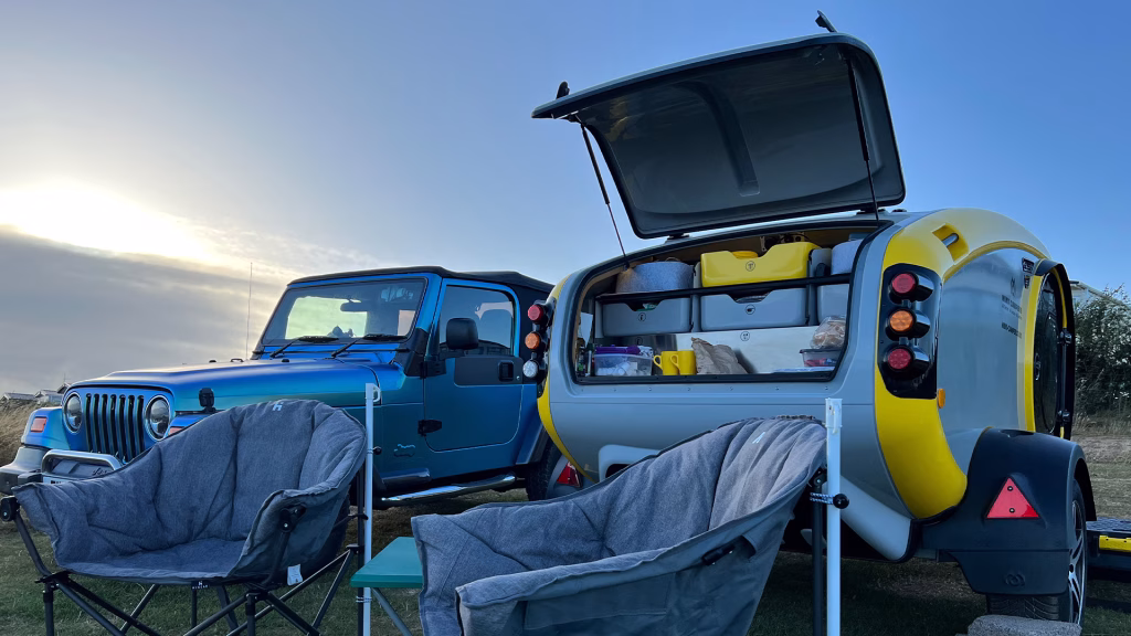 A MINK camper parked up on a UK campsite beside a blue Jeep Wrangler TJ. The sun is low and there are 2 collapsible camping chairs and a table in front of the MINK camper's open air kitchen.
