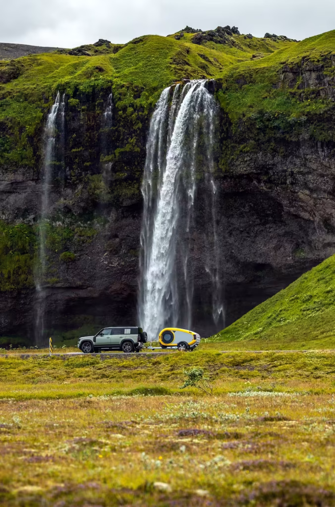 MINK camper passes in front of a waterfall, towed by a Landrover Defender.